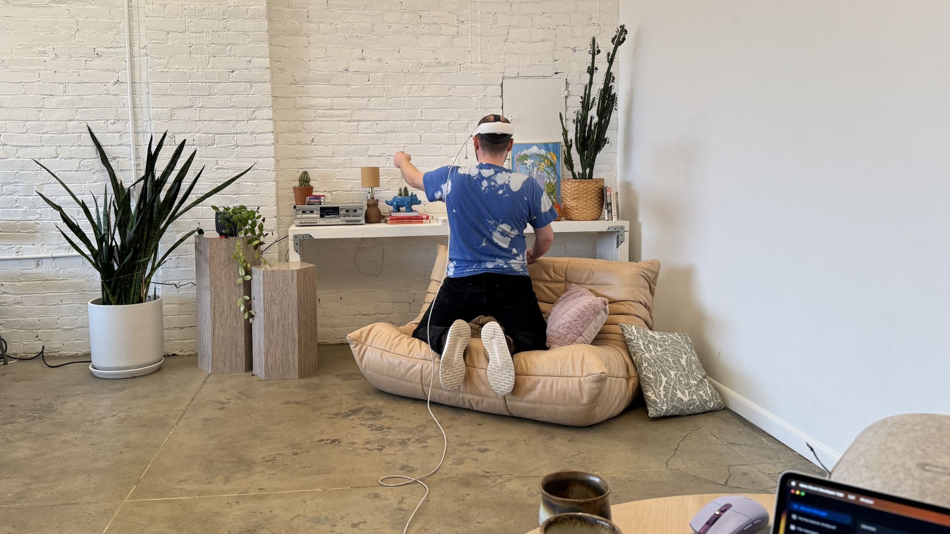 A man in a headset on his knees on a couch, facing away, leaning over a credenza, operating in virtual reality